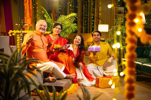 Ladus 'u kucaklayan ve birlikte gülümseyen Hintli aile, genç bir kızla birlikte, sari giymiş yaşlı bir anne, geleneksel giysili baba ve koca Diwali' yi kapalı mekanda kutluyor, çiçek rangoli ve kahkahalarla.