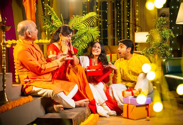 Diwali 'de altın kolye hediye eden Hintli genç kız sari içinde yaşlı anneyi şaşırtırken, geleneksel giyimli baba ve koca kapalı yerlerde diya, laddu ve çiçek rangoli dekoru ile gülümsüyor.