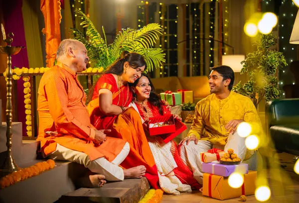 Diwali 'de altın kolye hediye eden Hintli genç kız sari içinde yaşlı anneyi şaşırtırken, geleneksel giyimli baba ve koca kapalı yerlerde diya, laddu ve çiçek rangoli dekoru ile gülümsüyor.