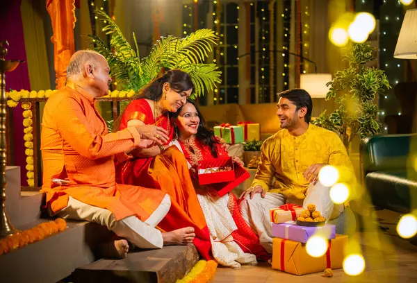Diwali 'de altın kolye hediye eden Hintli genç kız sari içinde yaşlı anneyi şaşırtırken, geleneksel giyimli baba ve koca kapalı yerlerde diya, laddu ve çiçek rangoli dekoru ile gülümsüyor.