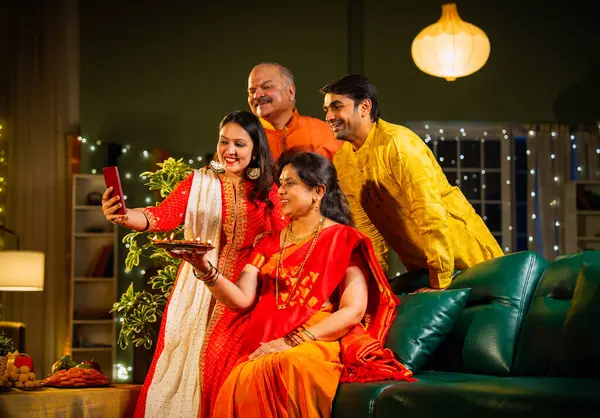 Diwali kutlaması sırasında selfie çeken, kıdemli ebeveynleri ve yetişkin çocukları olan Hintli aile akşam şenliklerinde birlikte oturan, ışıklarla süslenmiş, rahat koltukta mutlu bir şekilde gülümseyen.