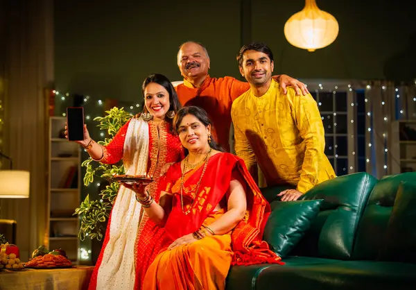 Kameraya boş akıllı telefon ekranı gösteren Hintli aile, genç oğlu ve kızı olan aile Diwali festivali sırasında rahat koltukta oturuyor, gülümsüyor ürün veya servisi tanıtıyor.