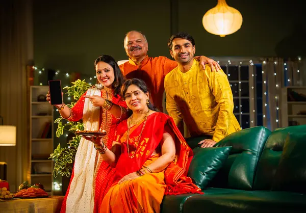 Kameraya boş akıllı telefon ekranı gösteren Hintli aile, genç oğlu ve kızı olan aile Diwali festivali sırasında rahat koltukta oturuyor, gülümsüyor ürün veya servisi tanıtıyor.
