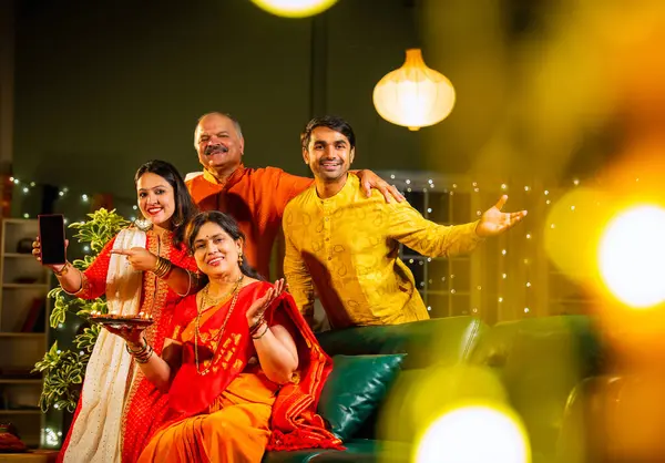 Kameraya boş akıllı telefon ekranı gösteren Hintli aile, genç oğlu ve kızı olan aile Diwali festivali sırasında rahat koltukta oturuyor, gülümsüyor ürün veya servisi tanıtıyor.