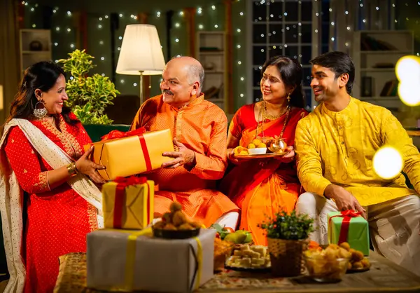 Yaşlı babaya Diwali hediyesi vermek, geleneksel giyimli Hintli kız festivalini evde kocası, annesi, laddu tabağı ve puja thali ile birlikte dekore edilmiş bir evde kanepede oturarak kutluyor.