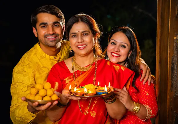 Puja thali ve laddu tabağıyla girişte duran, Hintli ailelere gülümseyen yaşlı aileler ve geleneksel şenlik elbisesi giyen genç çiftler süslü evde Hindu kutlamaları sırasında karşılanıyorlar.