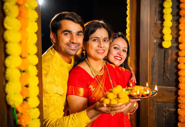 Puja thali ve laddu tabağıyla girişte duran, Hintli ailelere gülümseyen yaşlı aileler ve geleneksel şenlik elbisesi giyen genç çiftler süslü evde Hindu kutlamaları sırasında karşılanıyorlar.