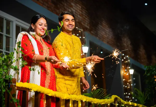 Kıvılcımlarla Diwali festivalinin tadını çıkaran geleneksel etnik kıyafetli genç Hintli çift, Diwali 'yi havai fişekler ve havai fişeklerle kutlarken süslü balkonda yan yana duruyorlar.