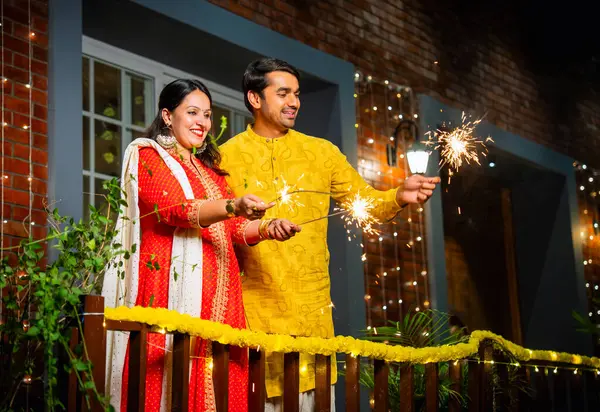 Kıvılcımlarla Diwali festivalinin tadını çıkaran geleneksel etnik kıyafetli genç Hintli çift, Diwali 'yi havai fişekler ve havai fişeklerle kutlarken süslü balkonda yan yana duruyorlar.