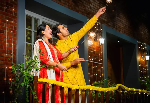 Kıvılcımlarla Diwali festivalinin tadını çıkaran geleneksel etnik kıyafetli genç Hintli çift, Diwali 'yi havai fişekler ve havai fişeklerle kutlarken süslü balkonda yan yana duruyorlar.