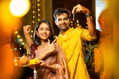 Gamalı haç çizerek ev anahtarlarını puja thali ile gösteriyorlar. Geleneksel Hintli genç çift fotoğraf makinelerini Hindu Diwali festivalindeki yeni evlerine girerken sevinçle selamlıyorlar.