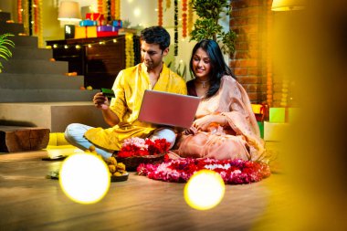 Diwali 'de çevrimiçi alışveriş yapan Hintli genç çift, çiçek ve ışıklarla süslenmiş şenlikli bir evde kanepe ve hediyelerle otururken bankamatik ya da kredi kartı kullanıyor.