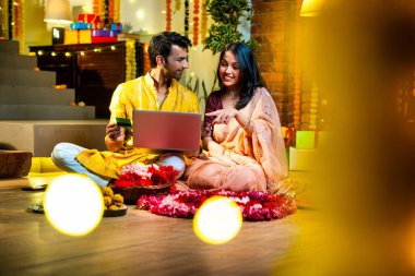 Diwali 'de çevrimiçi alışveriş yapan Hintli genç çift, çiçek ve ışıklarla süslenmiş şenlikli bir evde kanepe ve hediyelerle otururken bankamatik ya da kredi kartı kullanıyor.