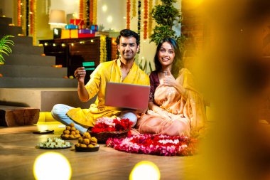 Diwali 'de çevrimiçi alışveriş yapan Hintli genç çift, çiçek ve ışıklarla süslenmiş şenlikli bir evde kanepe ve hediyelerle otururken bankamatik ya da kredi kartı kullanıyor.