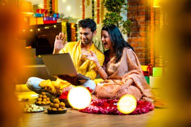 Diwali video konferansına katılan, etnik kökenli Hintli genç çift koltukta tatlı ve hediyelerle oturuyor, neşeli festival gecelerinde ışık ve çiçeklerle süslenmiş rahat bir evde akıllı telefon tutuyorlar.