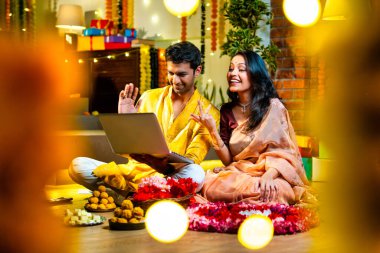 Diwali video konferansına katılan, etnik kökenli Hintli genç çift koltukta tatlı ve hediyelerle oturuyor, neşeli festival gecelerinde ışık ve çiçeklerle süslenmiş rahat bir evde akıllı telefon tutuyorlar.