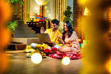 Diwali video konferansına katılan, etnik kökenli Hintli genç çift koltukta tatlı ve hediyelerle oturuyor, neşeli festival gecelerinde ışık ve çiçeklerle süslenmiş rahat bir evde akıllı telefon tutuyorlar.