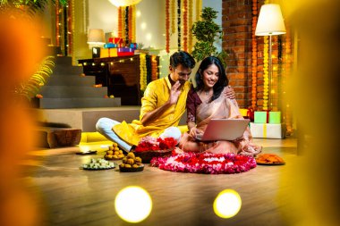 Diwali video konferansına katılan, etnik kökenli Hintli genç çift koltukta tatlı ve hediyelerle oturuyor, neşeli festival gecelerinde ışık ve çiçeklerle süslenmiş rahat bir evde akıllı telefon tutuyorlar.
