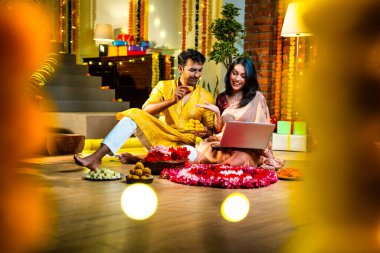 Diwali video konferansına katılan, etnik kökenli Hintli genç çift koltukta tatlı ve hediyelerle oturuyor, neşeli festival gecelerinde ışık ve çiçeklerle süslenmiş rahat bir evde akıllı telefon tutuyorlar.