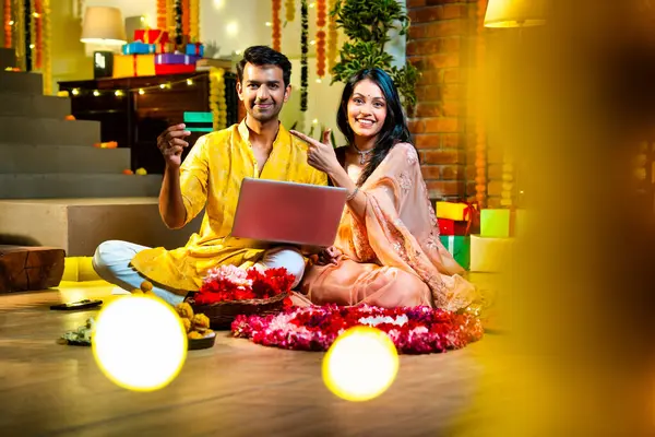 Diwali 'de çevrimiçi alışveriş yapan Hintli genç çift, çiçek ve ışıklarla süslenmiş şenlikli bir evde kanepe ve hediyelerle otururken bankamatik ya da kredi kartı kullanıyor.