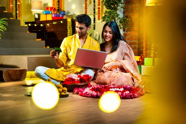 Diwali 'de çevrimiçi alışveriş yapan Hintli genç çift, çiçek ve ışıklarla süslenmiş şenlikli bir evde kanepe ve hediyelerle otururken bankamatik ya da kredi kartı kullanıyor.