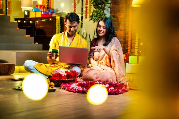 Diwali 'de çevrimiçi alışveriş yapan Hintli genç çift, çiçek ve ışıklarla süslenmiş şenlikli bir evde kanepe ve hediyelerle otururken bankamatik ya da kredi kartı kullanıyor.