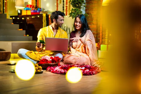 Diwali 'de çevrimiçi alışveriş yapan Hintli genç çift, çiçek ve ışıklarla süslenmiş şenlikli bir evde kanepe ve hediyelerle otururken bankamatik ya da kredi kartı kullanıyor.
