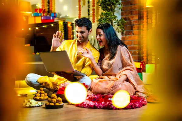 Diwali video konferansına katılan, etnik kökenli Hintli genç çift koltukta tatlı ve hediyelerle oturuyor, neşeli festival gecelerinde ışık ve çiçeklerle süslenmiş rahat bir evde akıllı telefon tutuyorlar.