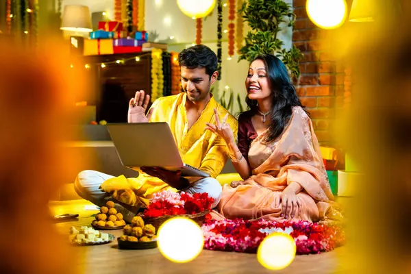 Diwali video konferansına katılan, etnik kökenli Hintli genç çift koltukta tatlı ve hediyelerle oturuyor, neşeli festival gecelerinde ışık ve çiçeklerle süslenmiş rahat bir evde akıllı telefon tutuyorlar.