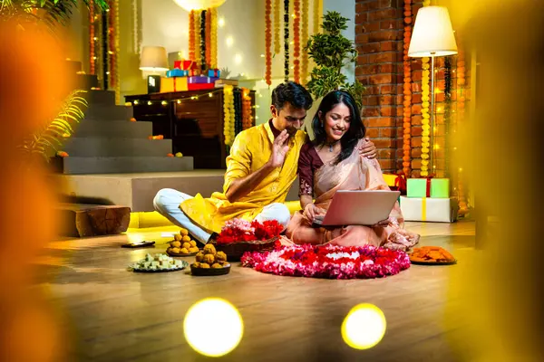 Diwali video konferansına katılan, etnik kökenli Hintli genç çift koltukta tatlı ve hediyelerle oturuyor, neşeli festival gecelerinde ışık ve çiçeklerle süslenmiş rahat bir evde akıllı telefon tutuyorlar.