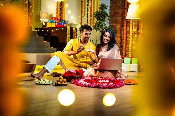 Diwali video konferansına katılan, etnik kökenli Hintli genç çift koltukta tatlı ve hediyelerle oturuyor, neşeli festival gecelerinde ışık ve çiçeklerle süslenmiş rahat bir evde akıllı telefon tutuyorlar.
