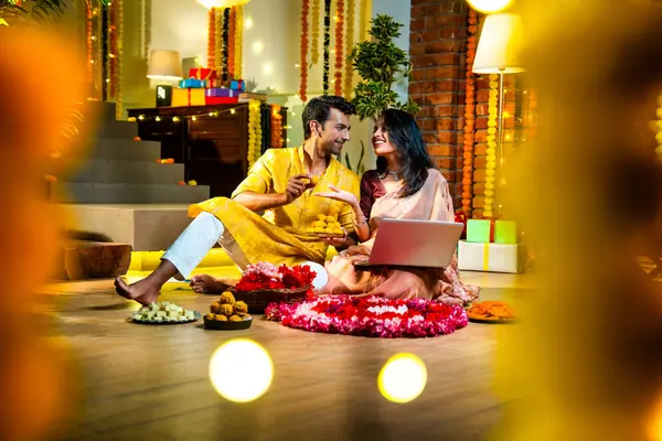 Diwali video konferansına katılan, etnik kökenli Hintli genç çift koltukta tatlı ve hediyelerle oturuyor, neşeli festival gecelerinde ışık ve çiçeklerle süslenmiş rahat bir evde akıllı telefon tutuyorlar.