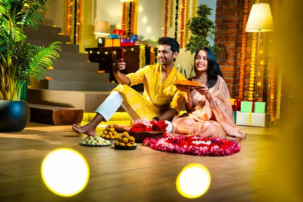 Arkadaşlarıyla Diwali 'de videoya çekilen, geleneksel giyinmiş Hintli genç çift, Deepavali gecesinde çiçek Rangoli' sinin yanında, süslenmiş evin içinde neşeli bir şenlik havasında kadife çiçekleri ve çelenklerle oturuyorlar.