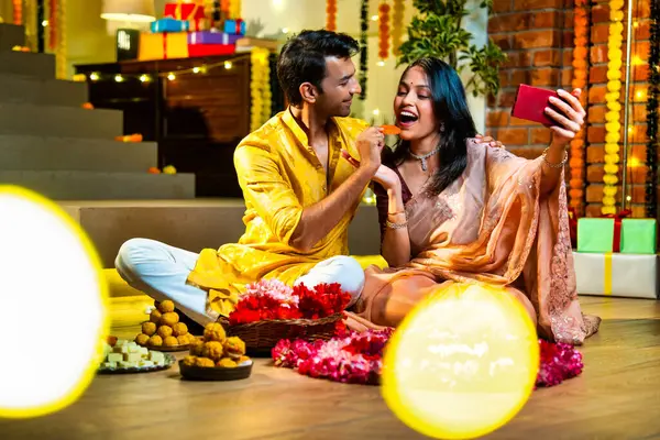 Arkadaşlarıyla Diwali 'de videoya çekilen, geleneksel giyinmiş Hintli genç çift, Deepavali gecesinde çiçek Rangoli' sinin yanında, süslenmiş evin içinde neşeli bir şenlik havasında kadife çiçekleri ve çelenklerle oturuyorlar.