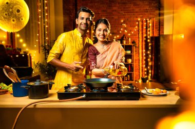 Diwali gecelerinde yemek pişirmek için tencereye yenilebilir yağ döken Hintli genç çift, aydınlatma ve çiçeklerle süslenmiş bir mutfakta birlikte yemek hazırlıyor ve neşeli bir havayla kutlama yapıyorlar.