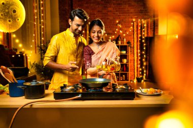 Diwali gecelerinde yemek pişirmek için tencereye yenilebilir yağ döken Hintli genç çift, aydınlatma ve çiçeklerle süslenmiş bir mutfakta birlikte yemek hazırlıyor ve neşeli bir havayla kutlama yapıyorlar.