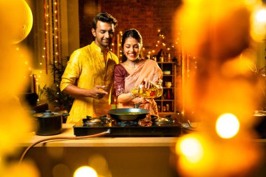 Diwali gecelerinde yemek pişirmek için tencereye yenilebilir yağ döken Hintli genç çift, aydınlatma ve çiçeklerle süslenmiş bir mutfakta birlikte yemek hazırlıyor ve neşeli bir havayla kutlama yapıyorlar.