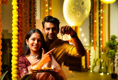 Ev anahtarını elinde tutan, etnik festivaldeki Hintli genç çift yeni evlerinde Diwali 'yi ilk kez kutluyor renkli çelenklerin önünde bayram gecesi neşe içinde duruyorlar.