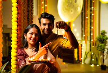 Ev anahtarını elinde tutan, etnik festivaldeki Hintli genç çift yeni evlerinde Diwali 'yi ilk kez kutluyor renkli çelenklerin önünde bayram gecesi neşe içinde duruyorlar.