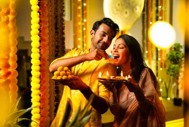 Hint çifti Hint yemeği ve laddu tabağı tutarak Diwali 'yi renkli çelenk süsleriyle kutluyor ve geceleri sıcak bir şekilde Mutlu Deepavali diliyor.