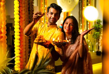 Hint çifti Hint yemeği ve laddu tabağı tutarak Diwali 'yi renkli çelenk süsleriyle kutluyor ve geceleri sıcak bir şekilde Mutlu Deepavali diliyor.