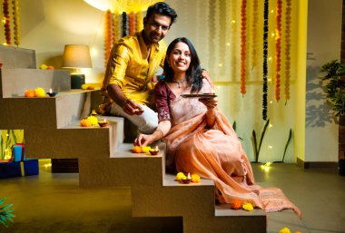 Diwali 'ye merdiven dayayan Hintli çift geleneksel şenlik kıyafetleri içinde geceleri Deepavali kutlamaları için teras lambaları yakıyor. Gülümsüyor, seviniyor ve renkli çle süsleniyor.