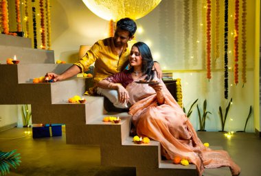 Diwali 'ye merdiven dayayan Hintli çift geleneksel şenlik kıyafetleri içinde geceleri Deepavali kutlamaları için teras lambaları yakıyor. Gülümsüyor, seviniyor ve renkli çle süsleniyor.