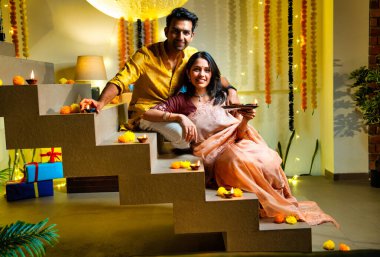 Diwali 'ye merdiven dayayan Hintli çift geleneksel şenlik kıyafetleri içinde geceleri Deepavali kutlamaları için teras lambaları yakıyor. Gülümsüyor, seviniyor ve renkli çle süsleniyor.