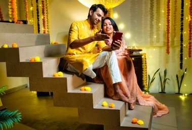 Merdivenlerdeki video görüşmelerine katılan Hintli çift Diwali gecesi kutlamalarında arkadaşlarıyla bağ kuruyor. Dekoratif merdivenlerde Divalar ve çelenklerle oturuyorlar.