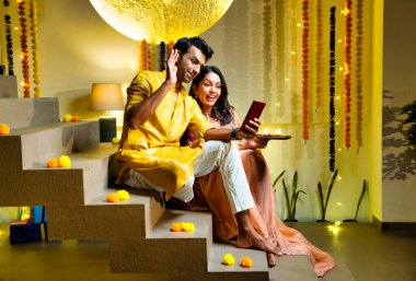 Merdivenlerdeki video görüşmelerine katılan Hintli çift Diwali gecesi kutlamalarında arkadaşlarıyla bağ kuruyor. Dekoratif merdivenlerde Divalar ve çelenklerle oturuyorlar.