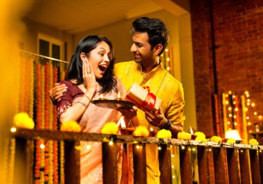 Karı koca Diwali kutlaması sırasında hediye veriyor, Hintli çift geleneksel kıyafet hediye ediyor ve evdeki ahşap korkuluklara gaz lambası yerleştiriyor, parıltılı diya ve dekorasyonlarla şenlikli bir akşamın tadını çıkarıyor.