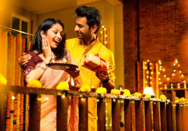 Karı koca Diwali kutlaması sırasında hediye veriyor, Hintli çift geleneksel kıyafet hediye ediyor ve evdeki ahşap korkuluklara gaz lambası yerleştiriyor, parıltılı diya ve dekorasyonlarla şenlikli bir akşamın tadını çıkarıyor.