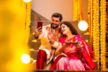 Diwali 'ye video çağrısı yapan çift, Saree' de Hintli Genç Evli Çifti ve Şenlik Gecesinde Smartphone aracılığıyla Akrabalarla Gülümseyen Diya Thali 'yi tutan süslü merdivenlerde oturan Kurta