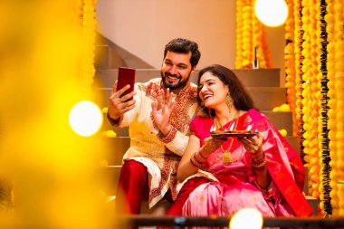 Diwali 'ye video çağrısı yapan çift, Saree' de Hintli Genç Evli Çifti ve Şenlik Gecesinde Smartphone aracılığıyla Akrabalarla Gülümseyen Diya Thali 'yi tutan süslü merdivenlerde oturan Kurta